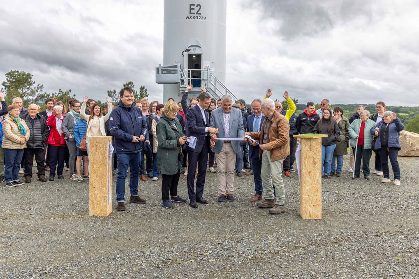 A crowd gathers at a wind turbine site, with several people engaging in a ribbon-cutting ceremony amidst cloudy weather.