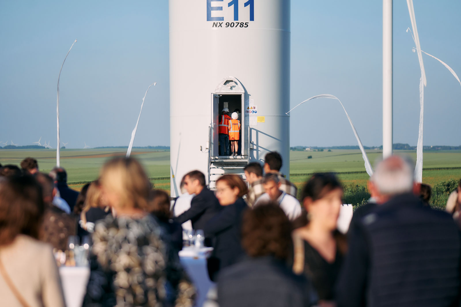 A crowd gathers near a wind turbine with two workers preparing to ascend. The landscape features green fields and a clear sky.
