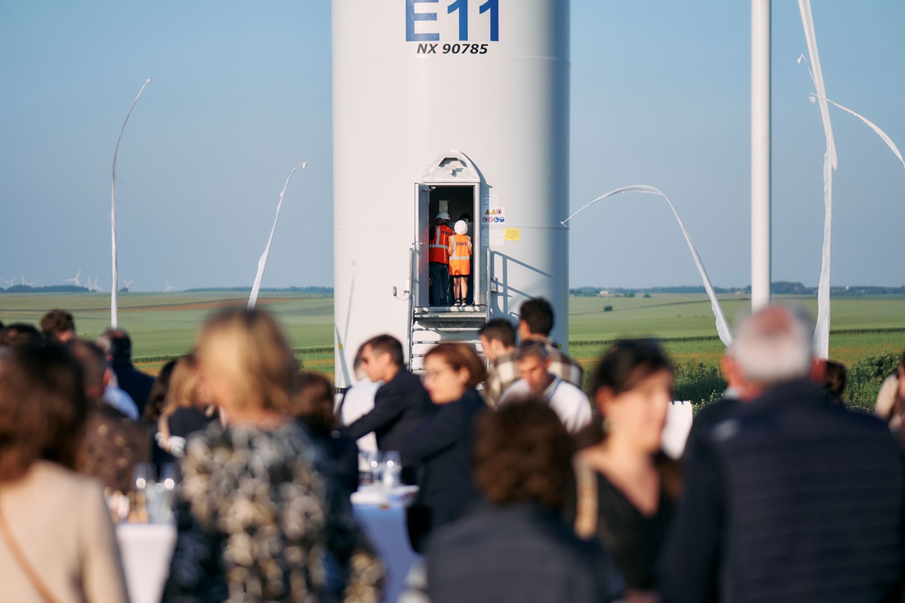 A crowd gathers near a wind turbine with two workers preparing to ascend. The landscape features green fields and a clear sky.