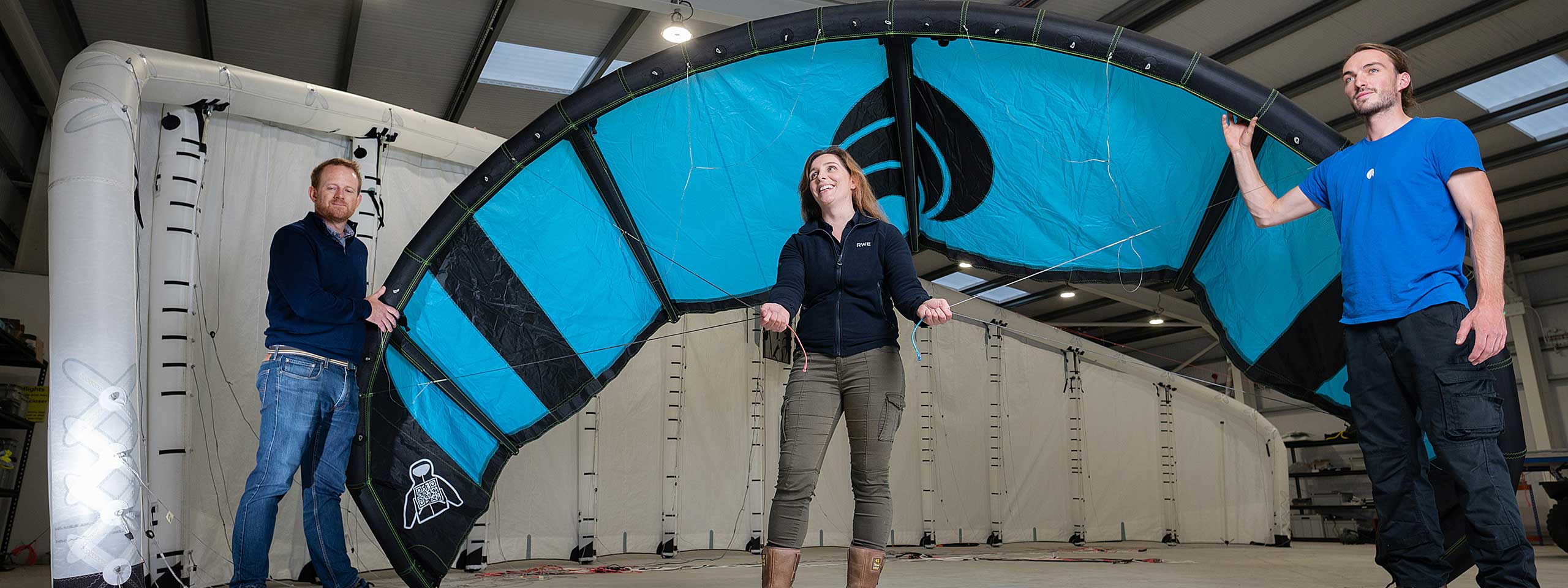 Drei Personen im Innenraum halten einen Kitesurf-Schirm. Der Schirm ist schwarz und türkis mit einem auffälligen Design.