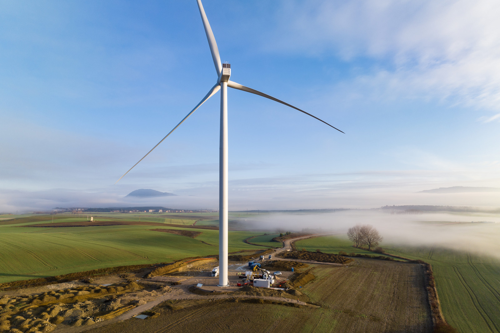 A tall wind turbine stands on a green field, surrounded by fog and fields extending to distant mountains.