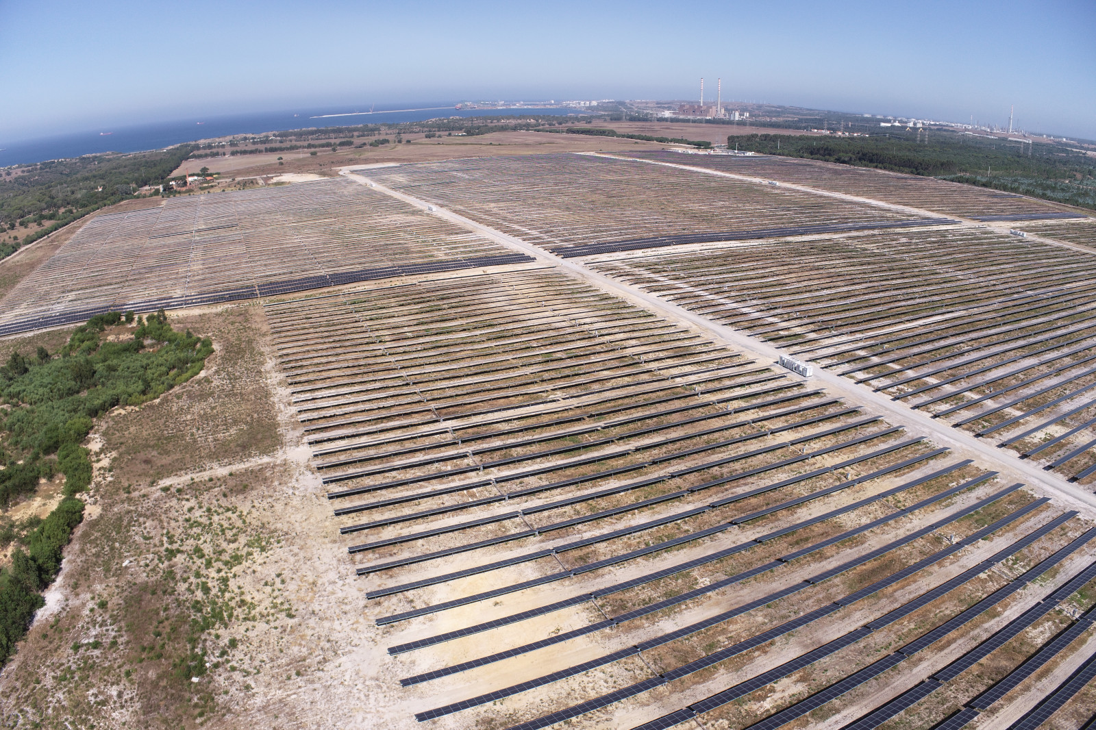 Aerial view of a vast solar farm with numerous panels, bordered by greenery and the sea in the distance.