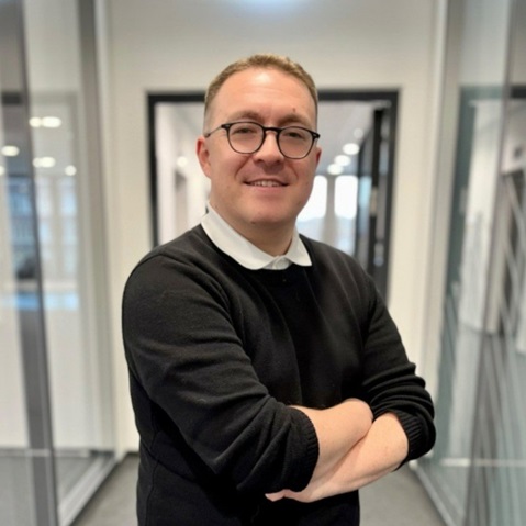 A person in an office with crossed arms, wearing a black jumper with a white collar. Portrait of Lee Clark.
