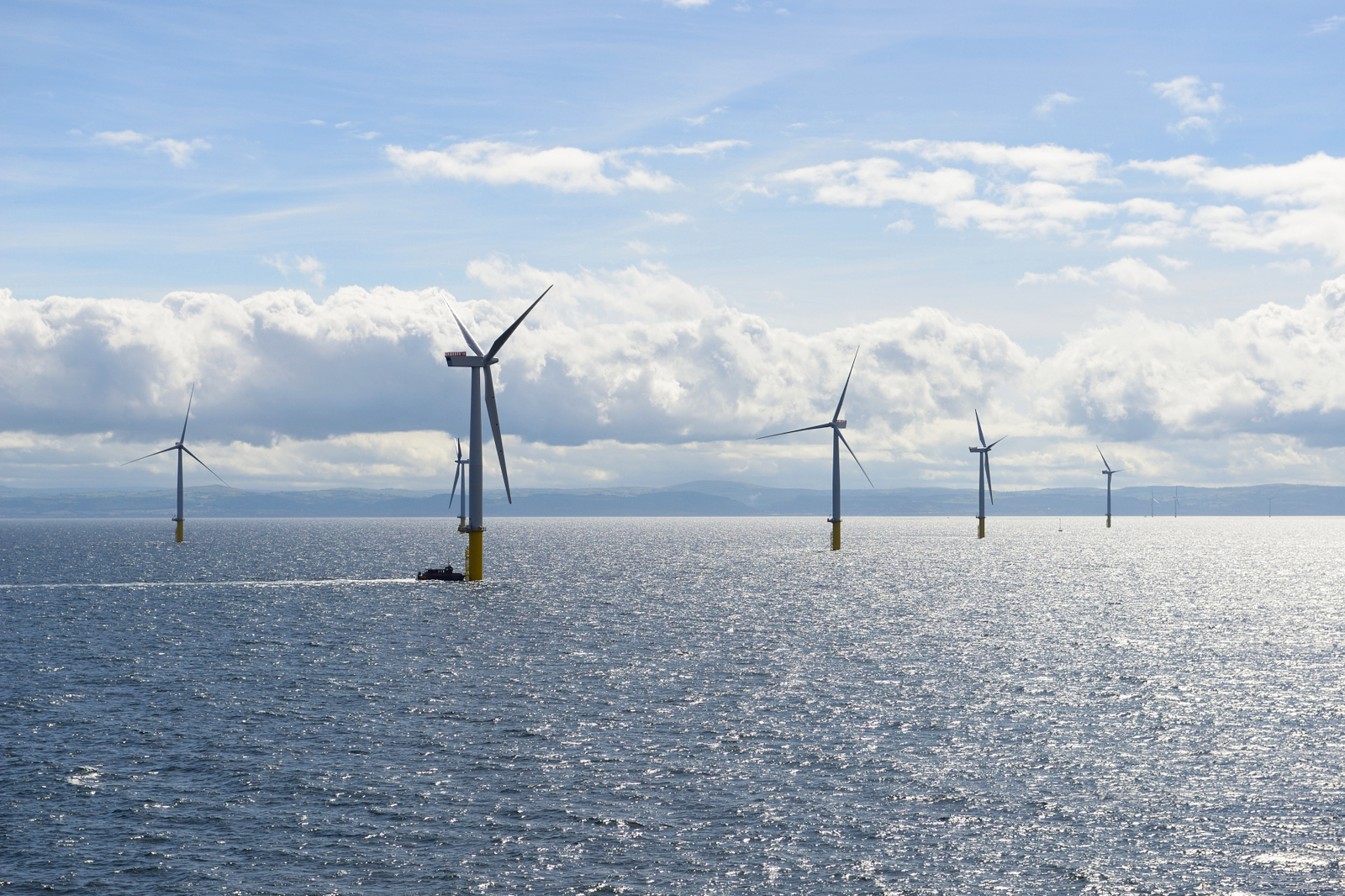 White offshore wind turbines with yellow bases stand in a sunlit blue-gray sea under a partly cloudy sky reflecting bright highlights.