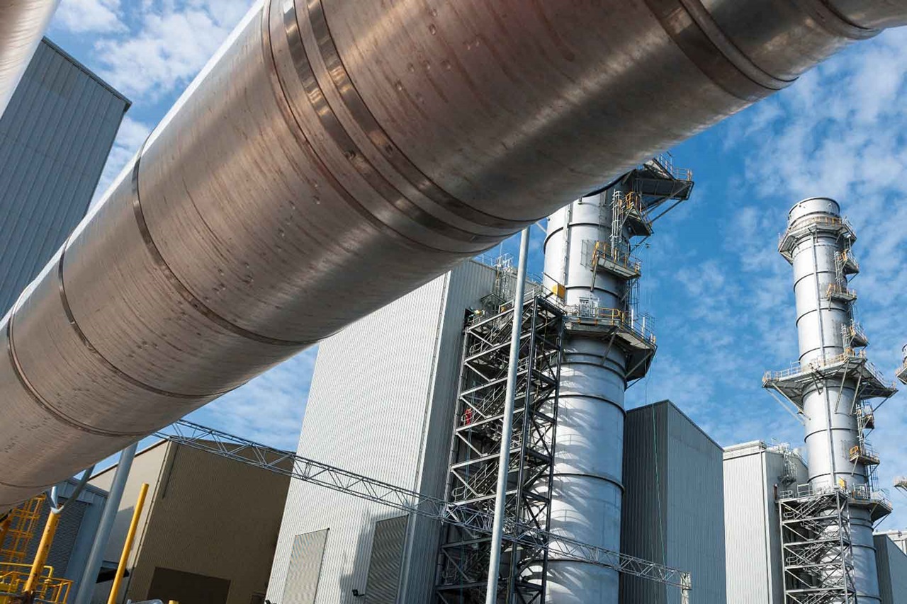 Eine Nahaufnahme von großen Stahlrohren an einem Industriestandort mit sichtbaren Rauchschornsteinen unter einem blauen Himmel.