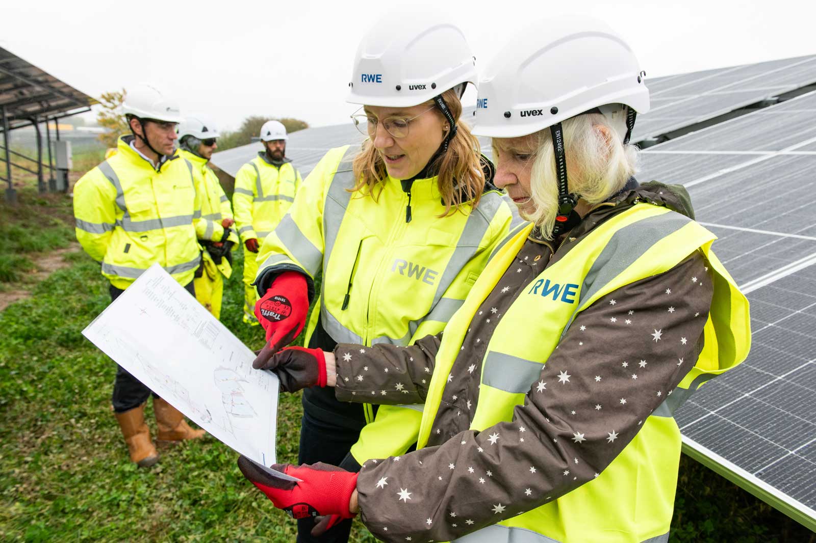 Two individuals in safety gear examine a plan near solar panels, with others in the background observing the activity.