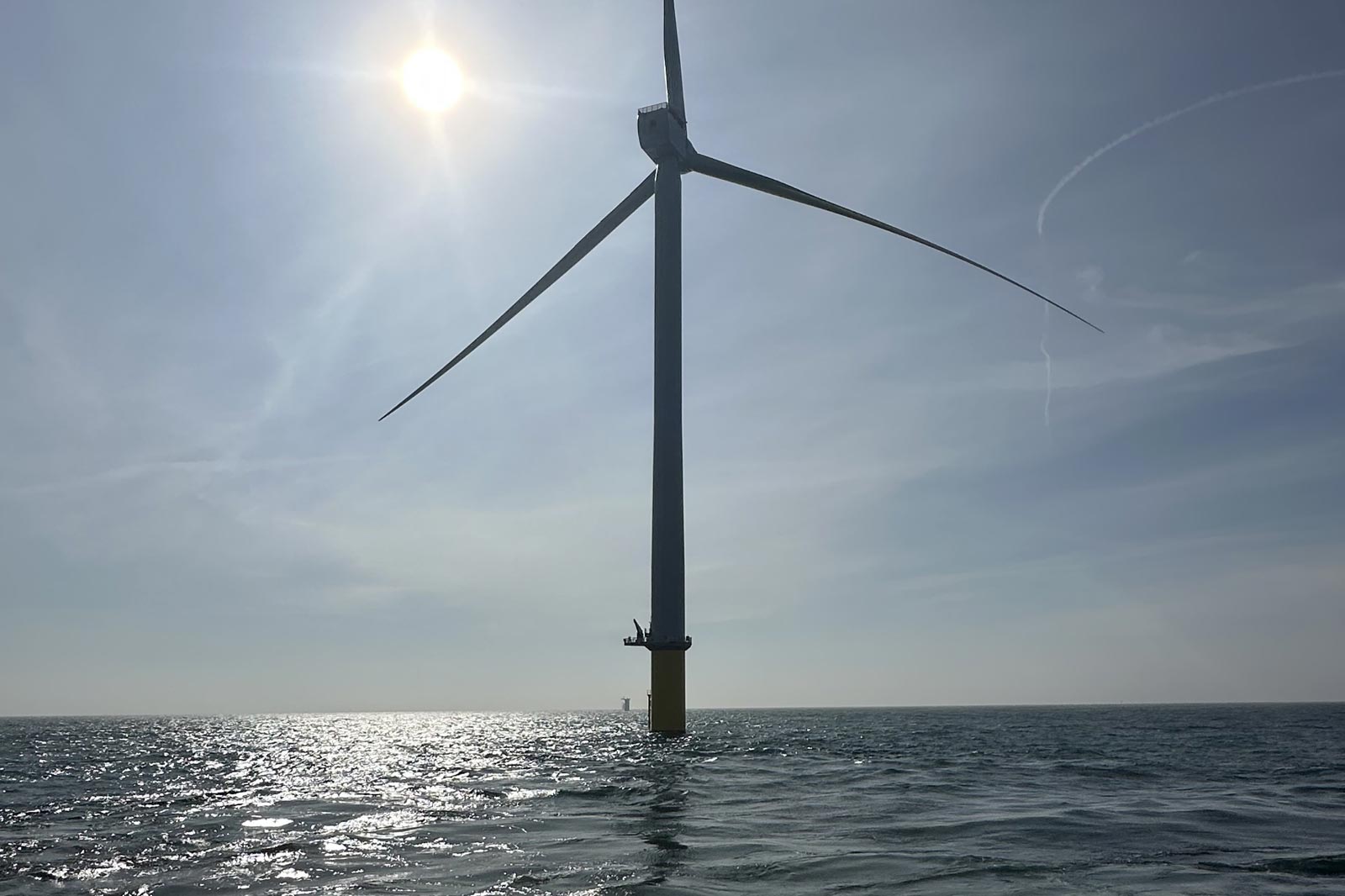 A tall offshore wind turbine stands in the sea, with a bright sun overhead against a clear sky.