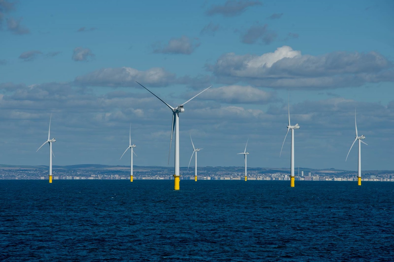 A row of wind turbines on the water with yellow bases against a clear sky and a city skyline in the background.