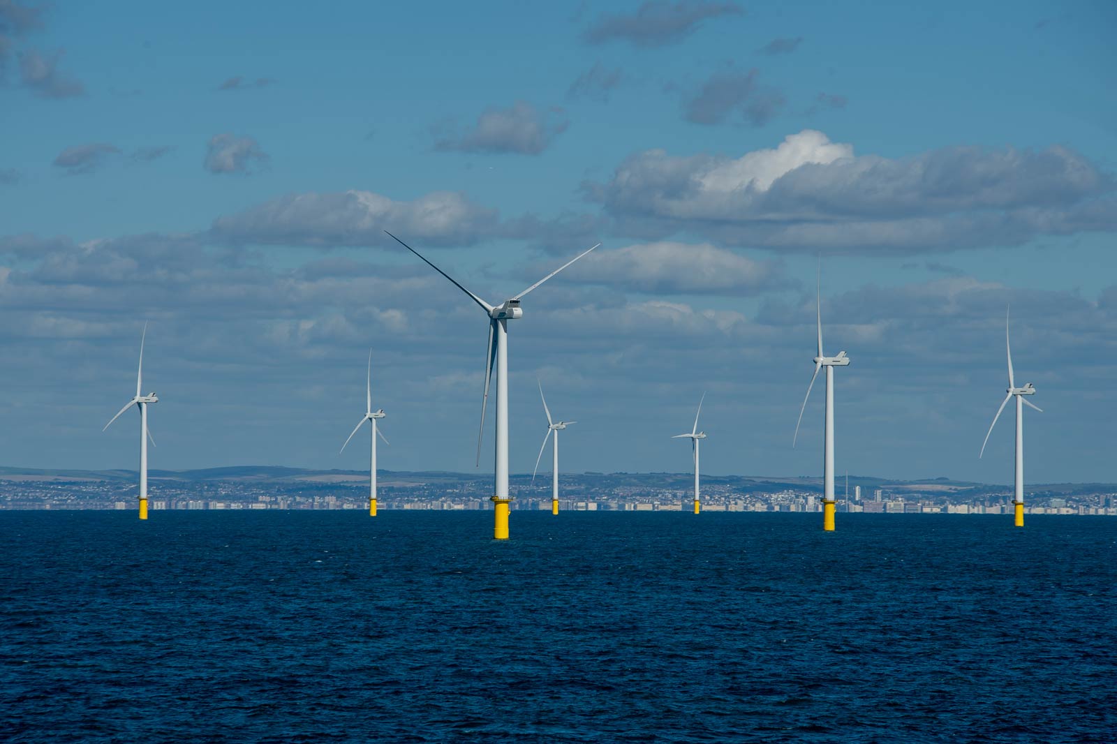 A row of wind turbines on the water with yellow bases against a clear sky and a city skyline in the background.