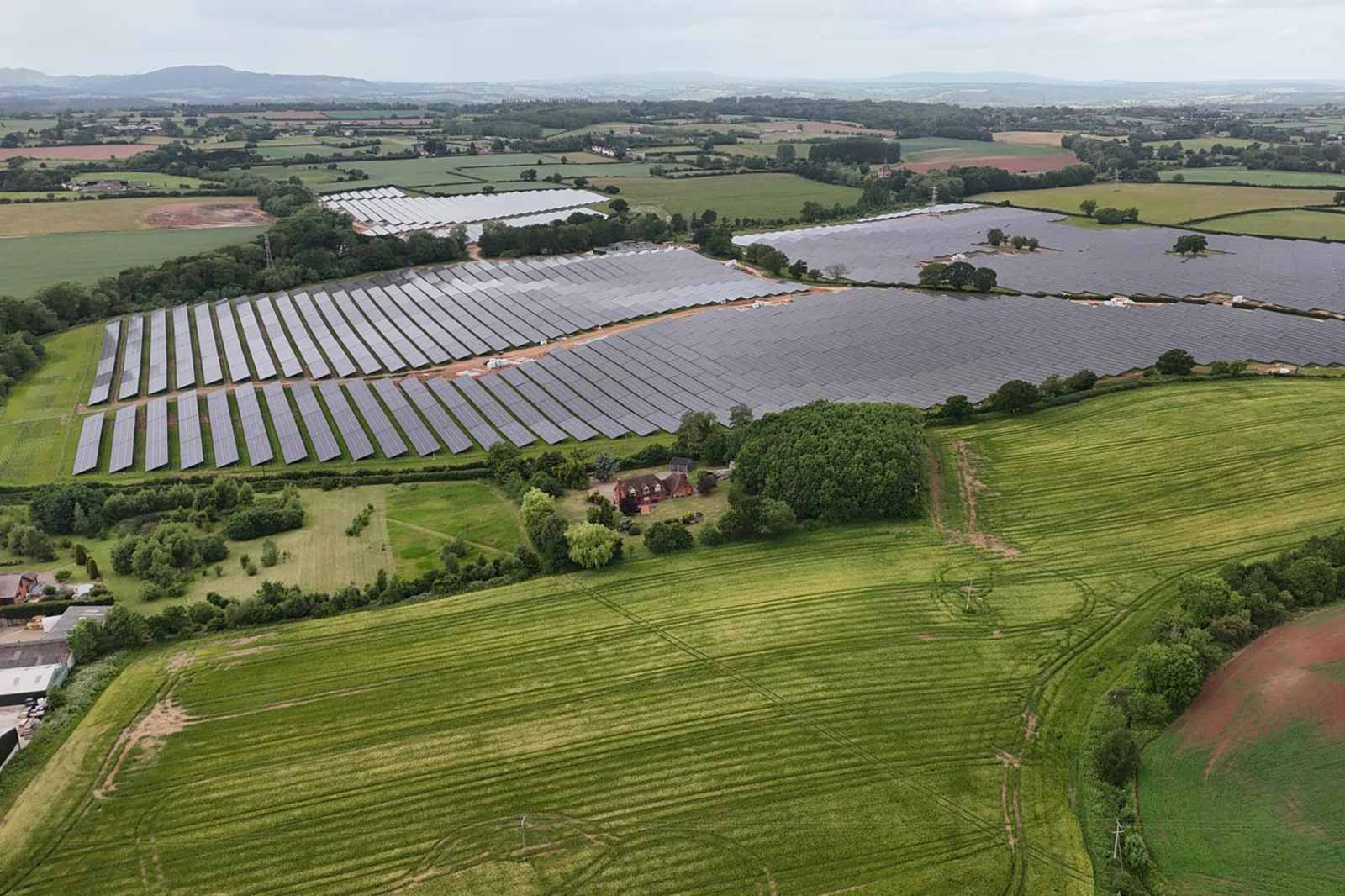 Aerial view of a solar farm surrounded by green fields, with a small house and trees in the foreground.
