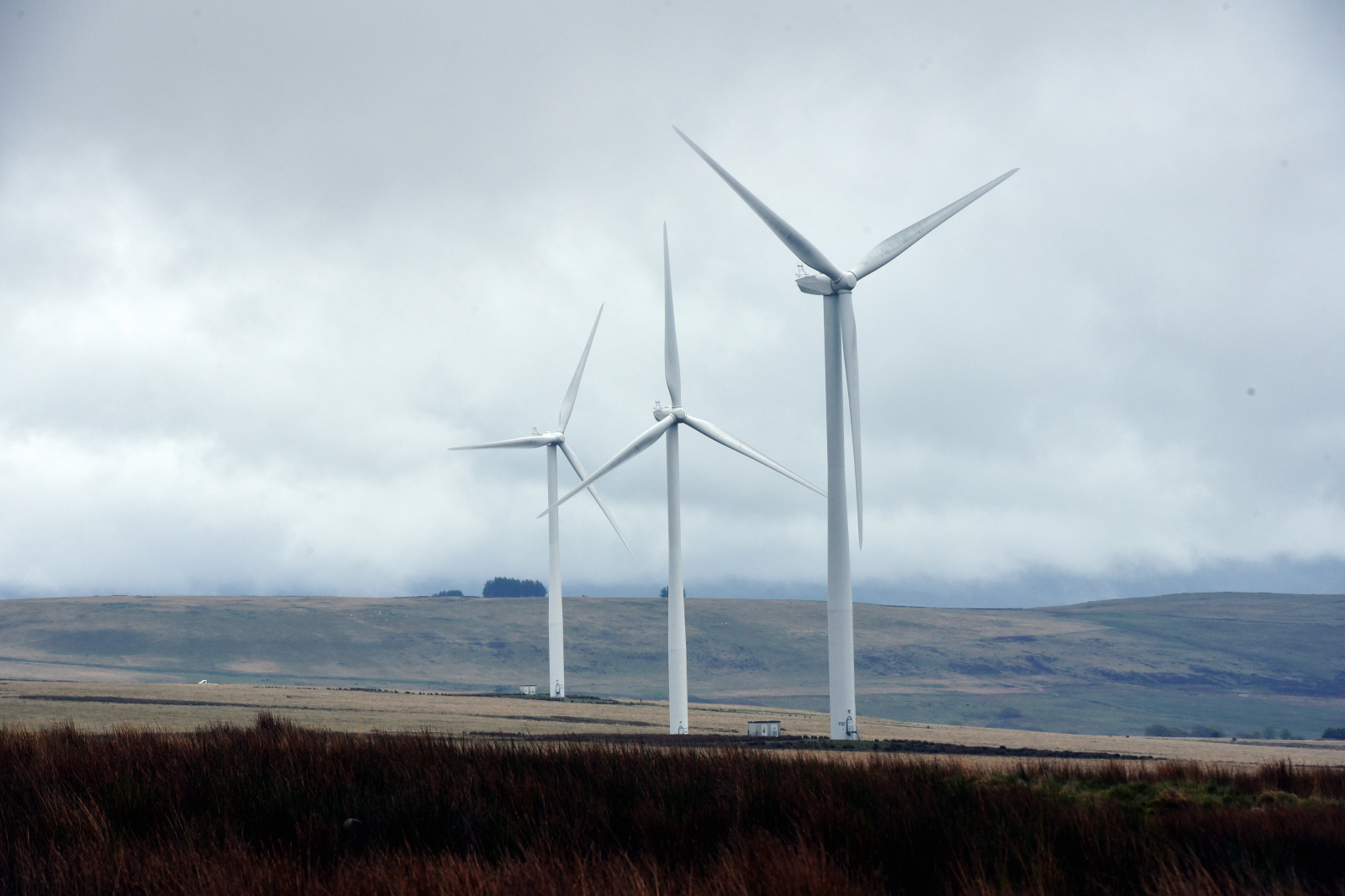 Three tall wind turbines stand against a cloudy sky in a rural landscape, surrounded by grassy fields and distant hills.