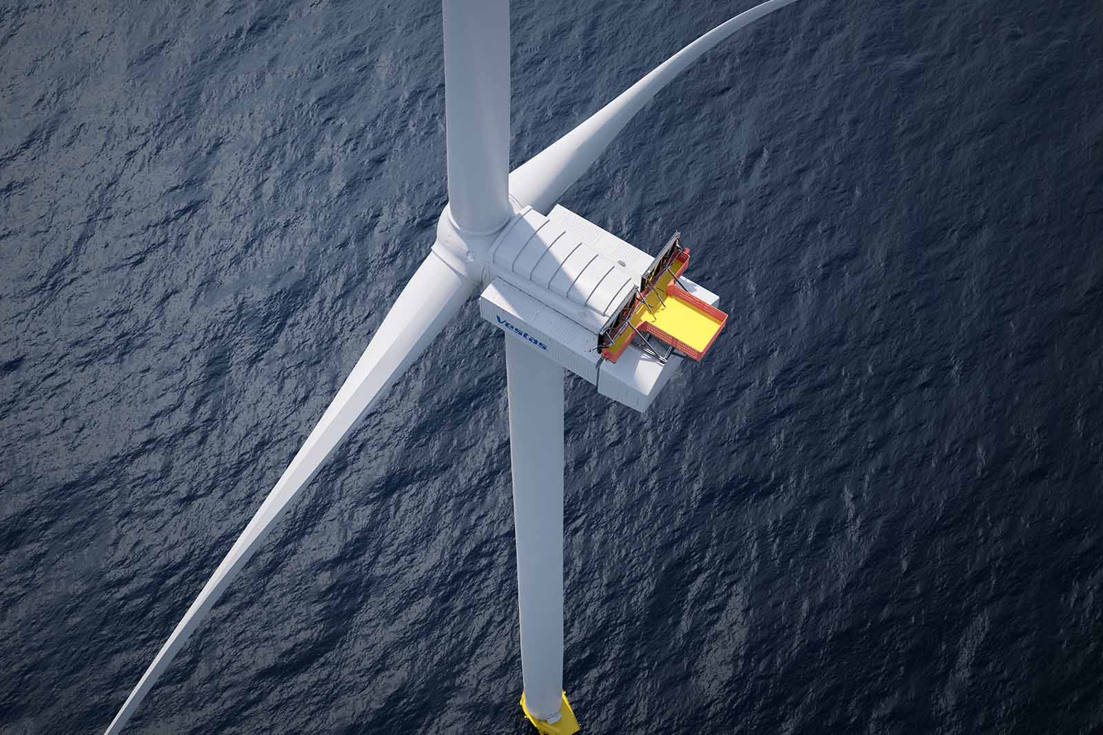 An aerial view of an offshore wind turbine with three white blades, a white nacelle and a yellow maintenance platform above dark sea.