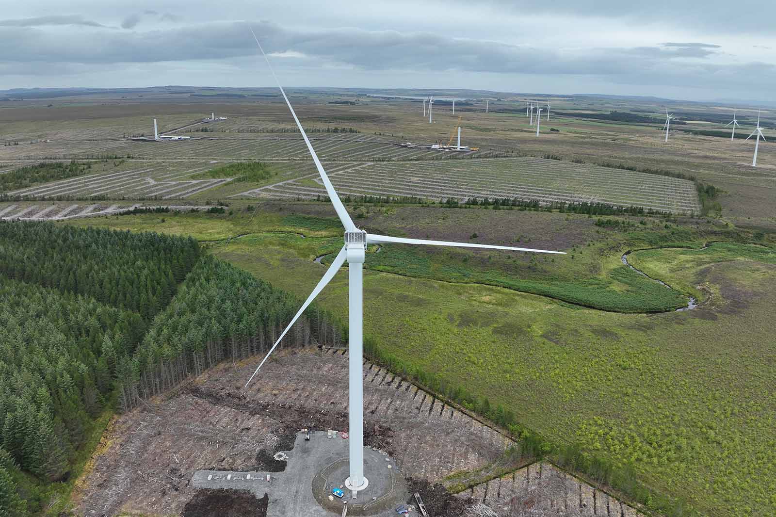 A white three-bladed wind turbine stands on a concrete base in a cleared brown patch amid green fields and distant turbines under a gray sky.
