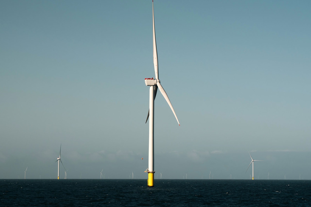 A large white offshore wind turbine with a yellow cylindrical base rises from a dark blue sea under a pale blue sky.