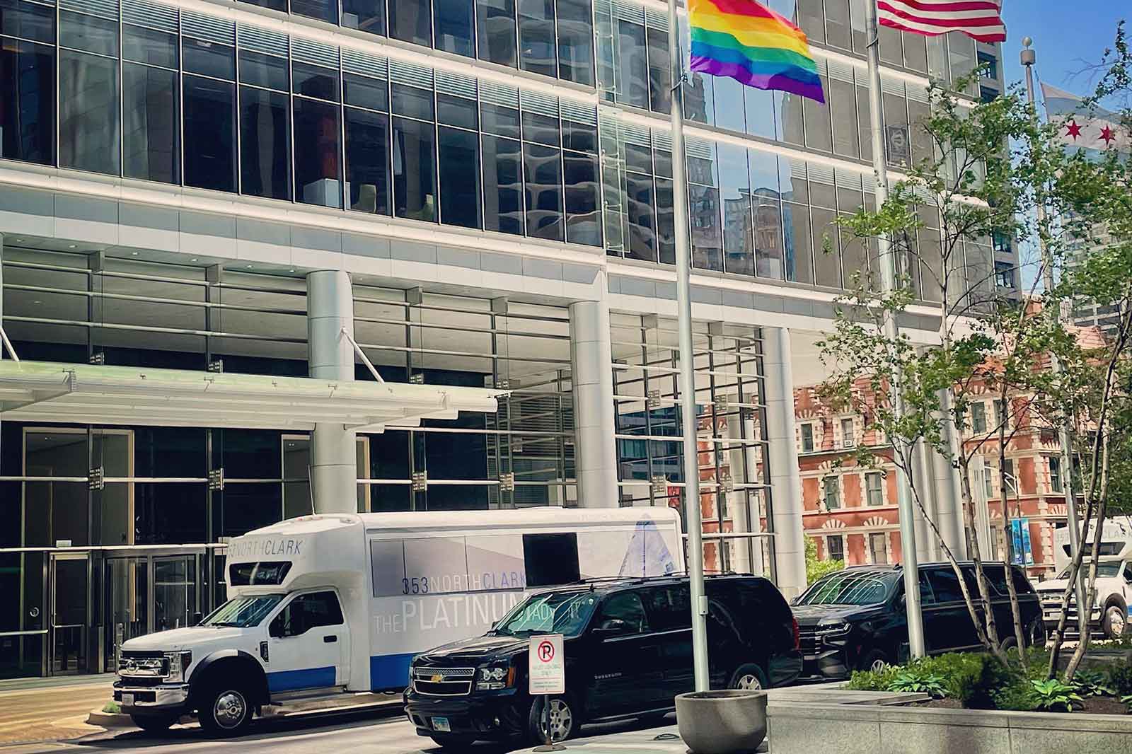 A modern building with large glass windows, featuring a rainbow flag and two American flags. Vehicles are parked outside.