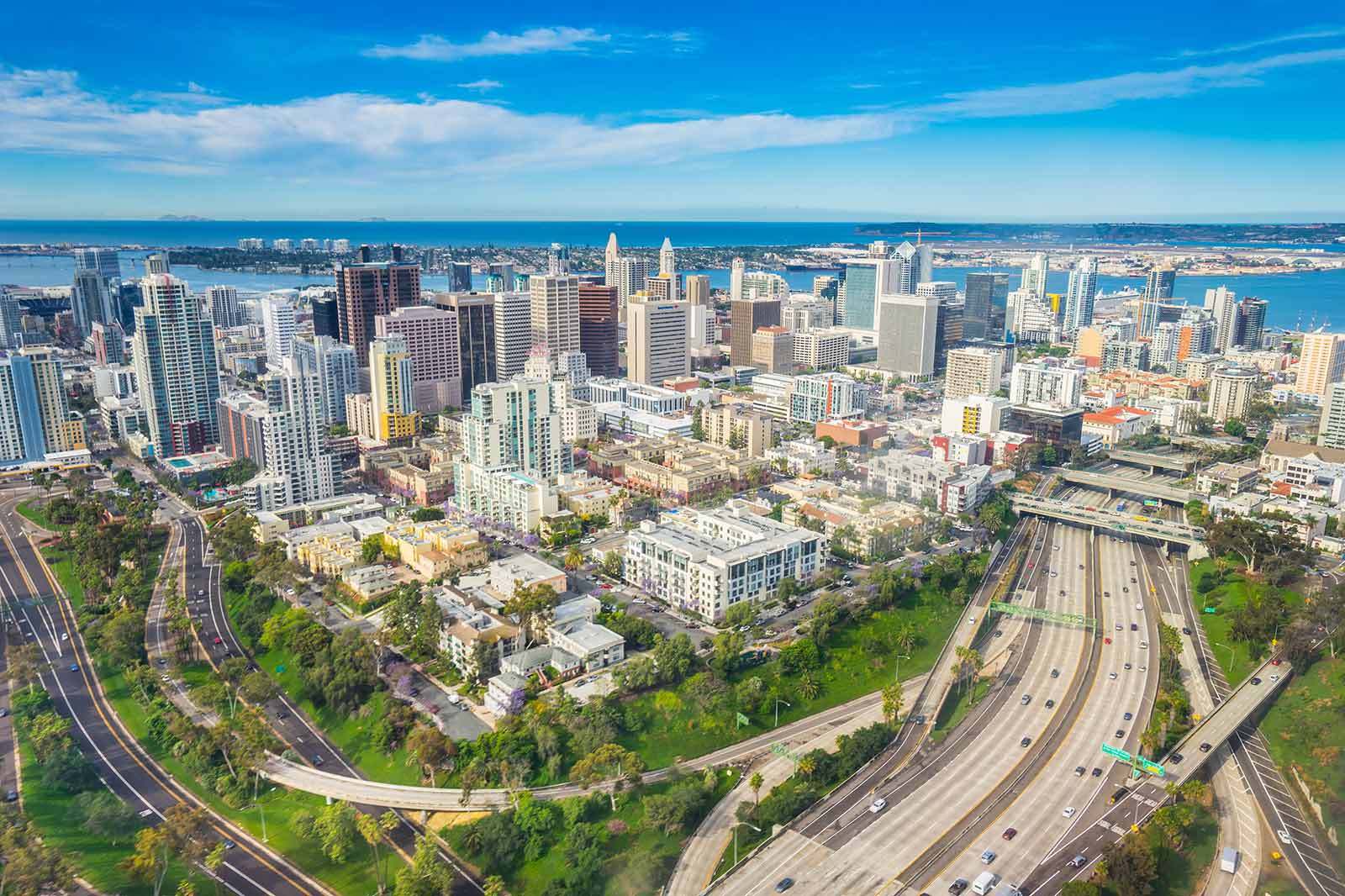 Aerial view of a vibrant city skyline with tall buildings, greenery, and busy highways leading to the ocean.