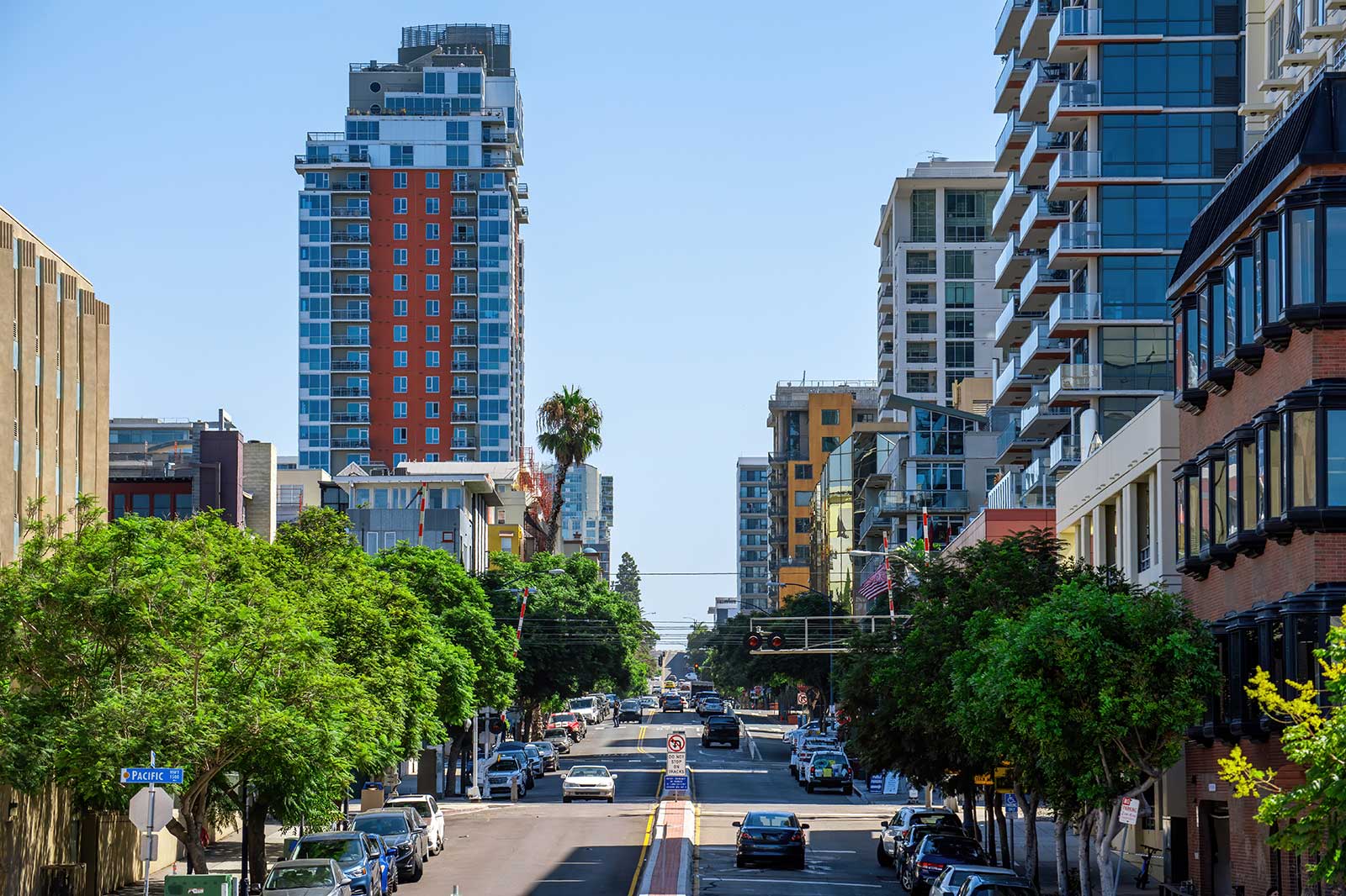 A vibrant urban street lined with modern buildings, lush trees, and parked cars, under a clear blue sky.