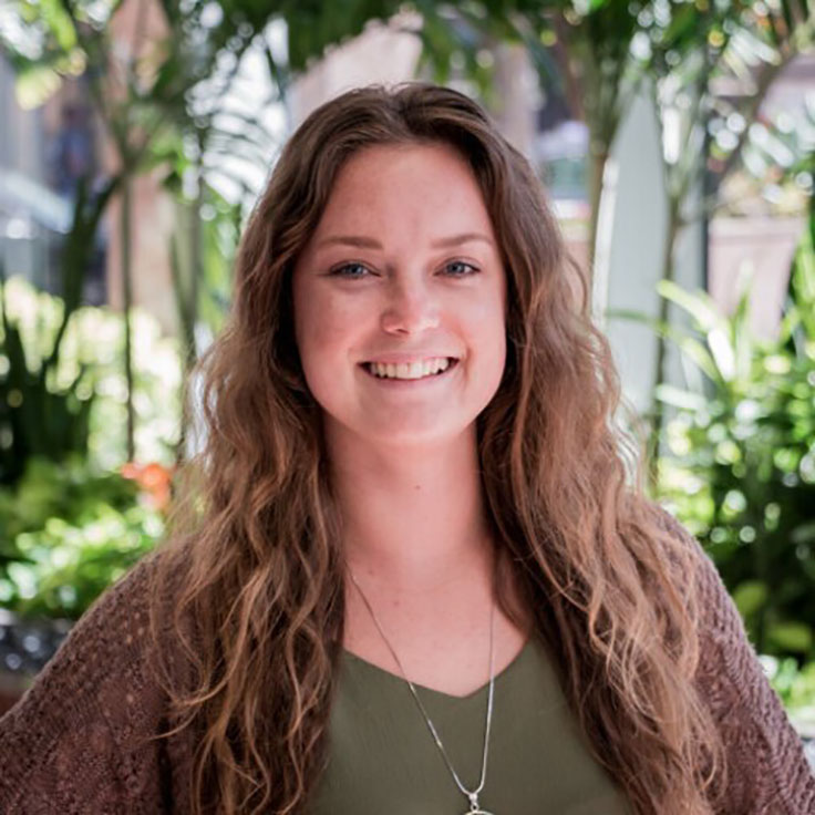 A person with long, wavy hair stands outdoors among lush greenery, wearing a green top and a knitted cardigan.