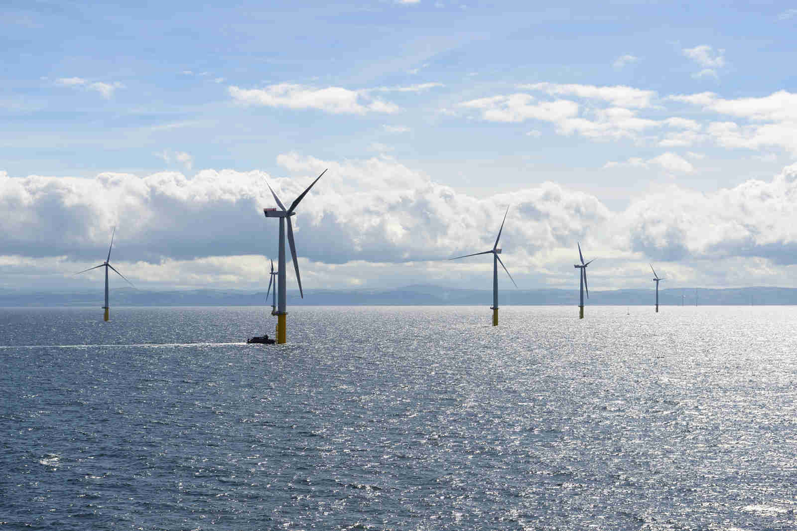 A scenic view of offshore wind turbines in the ocean, with a boat travelling between them under a cloudy sky.