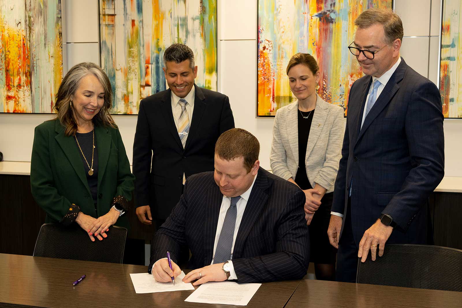 A group of professionals gathered around a table as one person signs a document, with colourful abstract paintings in the background.