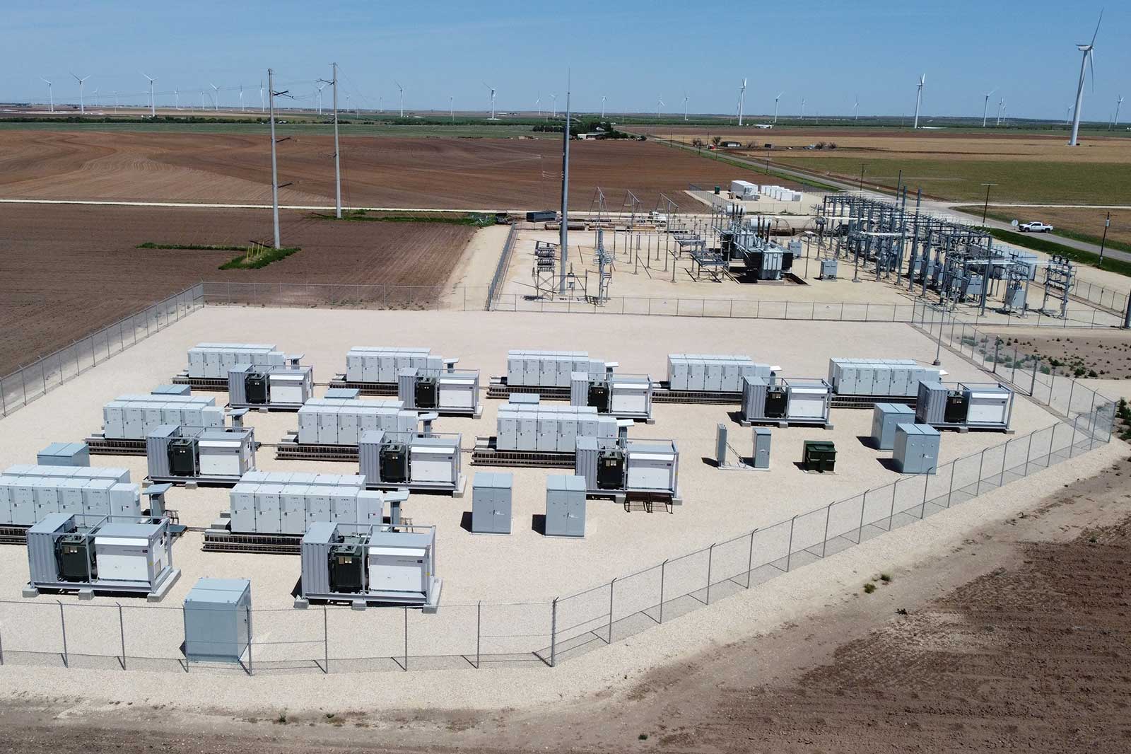 Aerial view of an energy facility with battery storage units, power lines, and wind turbines in the background.