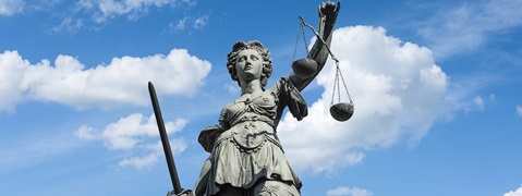 Eine Statue der Justitia mit Schwert und Waage vor einem blauen Himmel mit Wolken.