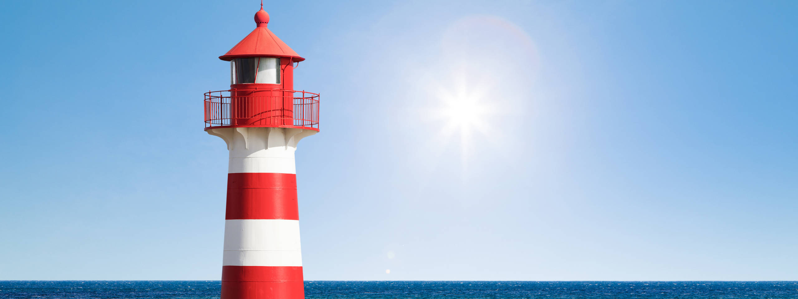 Ein rot-weiß gestreifter Leuchtturm steht am Ozean unter einem klaren blauen Himmel, während die Sonne hell scheint.