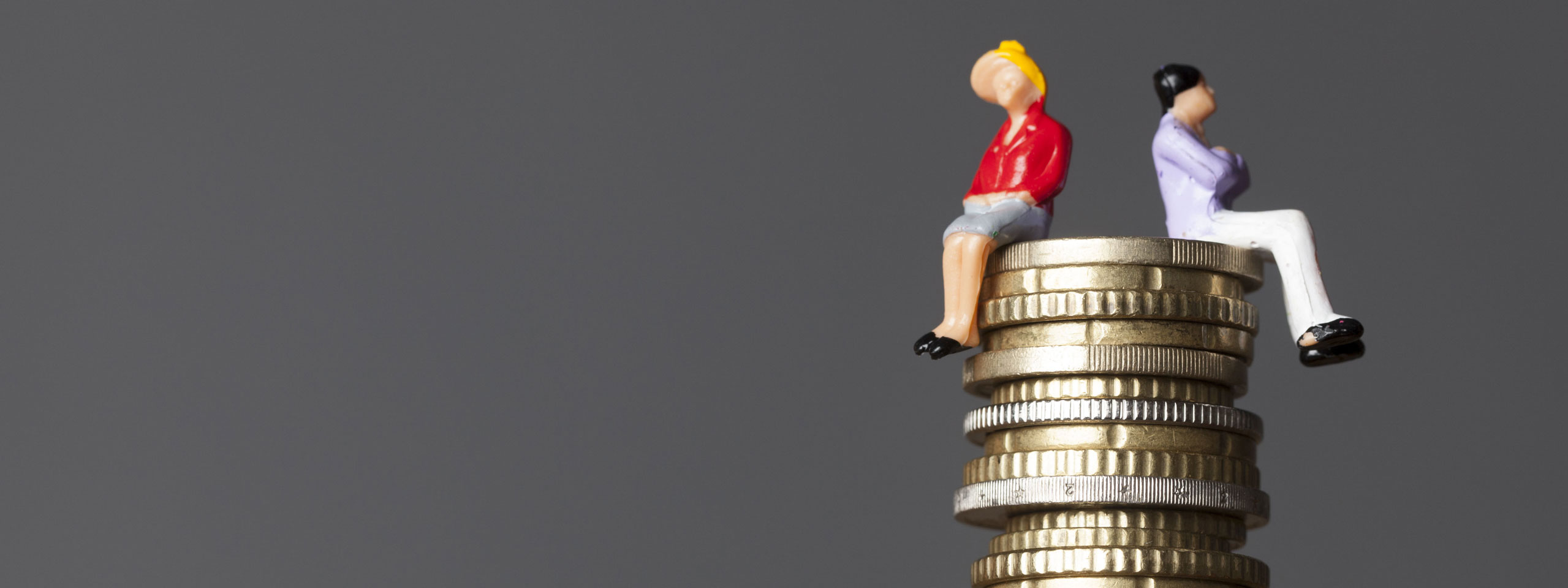 Two miniature figures sit atop a stack of coins against a grey background, symbolising financial themes.