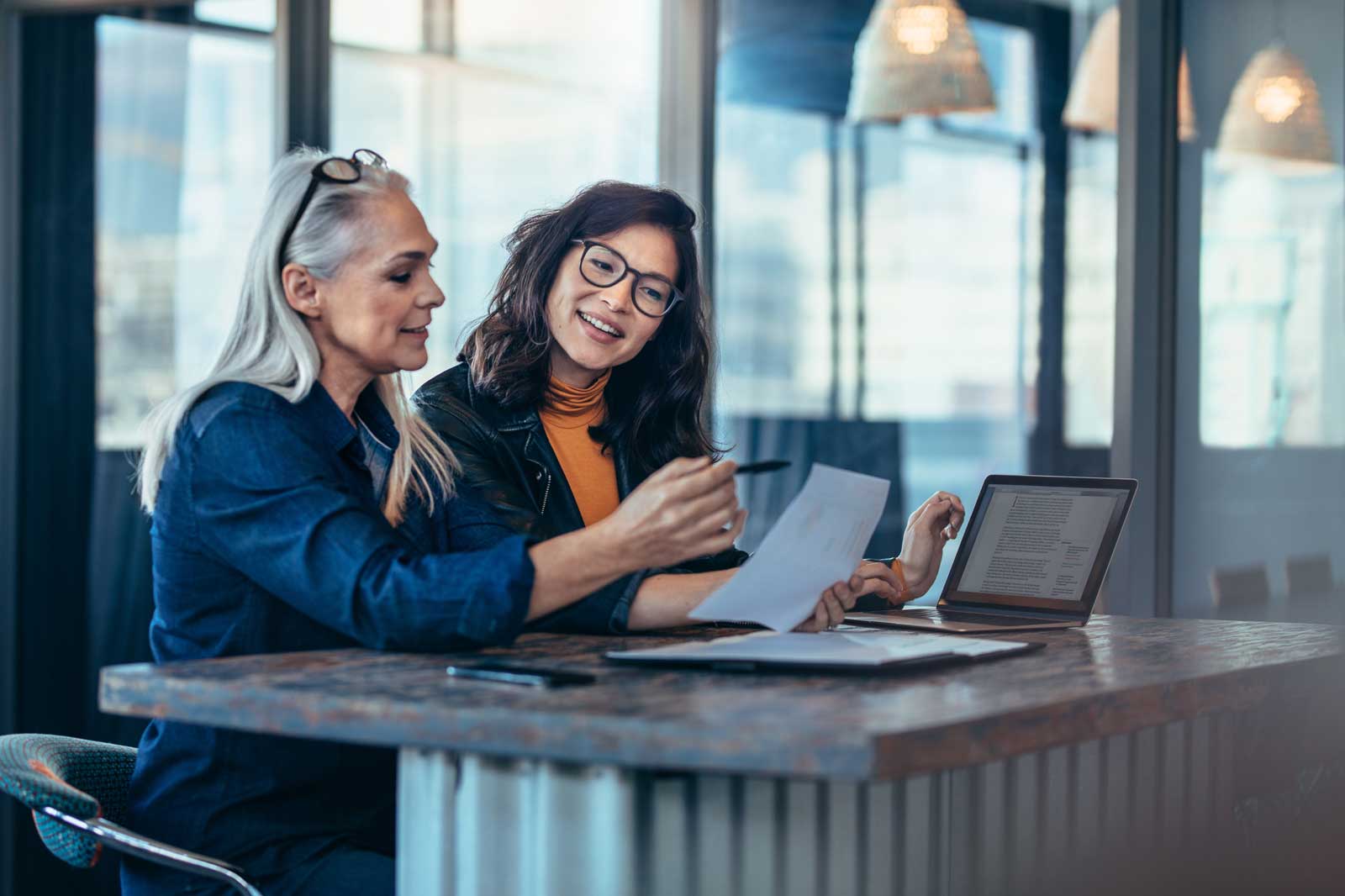 Two women sit at a table discussing while using documents and a laptop.