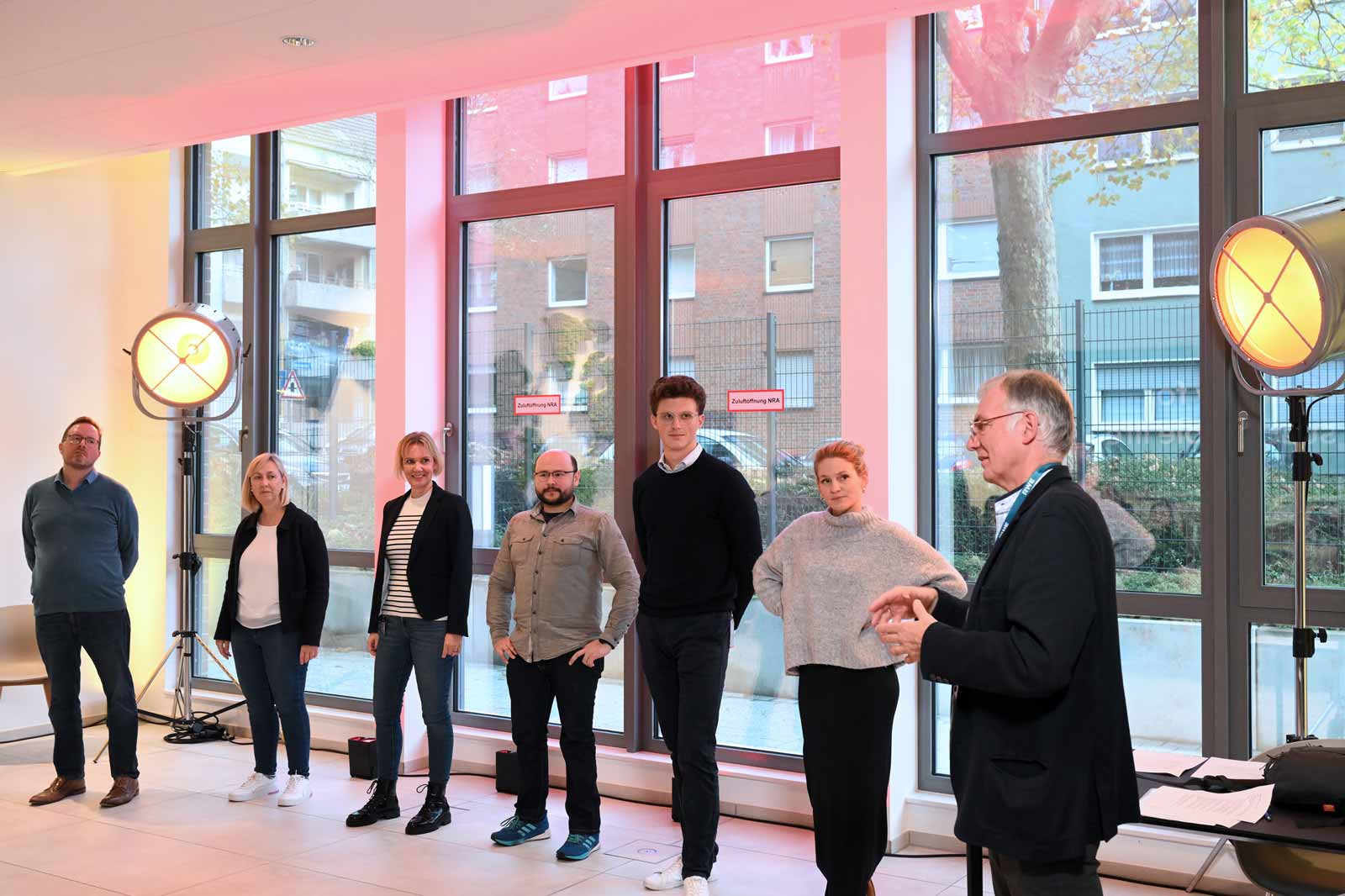 A group of people stands in a bright room with large windows and spotlights.