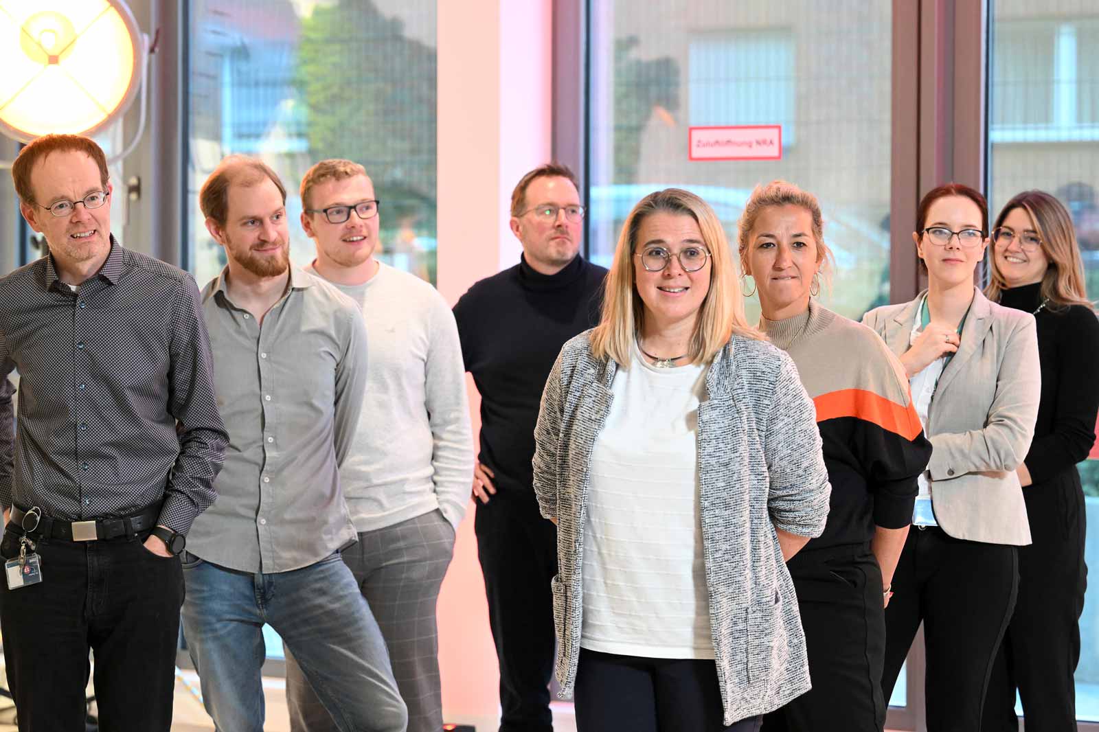 A group of people stands in a bright room with large windows. The individuals are wearing casual clothing.