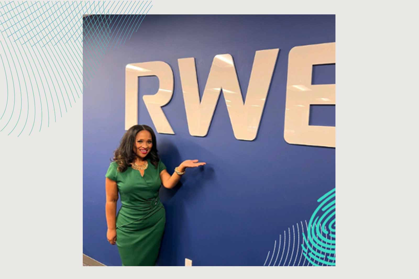 A person in a green dress poses beside a large 'RWE' sign against a blue wall. Portrait of Natasha Bowmann.