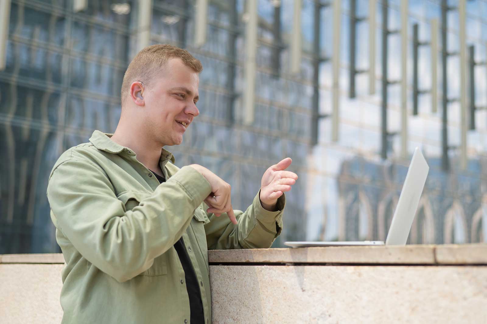 Eine Person gestikuliert beim Sprechen an einem Laptop im Freien, vor einem modernen Glasgebäude im Hintergrund.