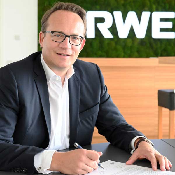 Ein Mann in einem schwarzen Anzug sitzt an einem Tisch, während er ein Dokument unterschreibt. Im Hintergrund ist das Logo von RWE zu sehen. Portrait von Dr. Markus Krebber.