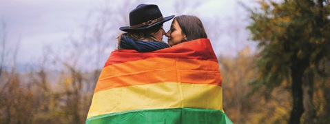 A couple embraces while wrapped in a rainbow flag, set against an autumn landscape.
