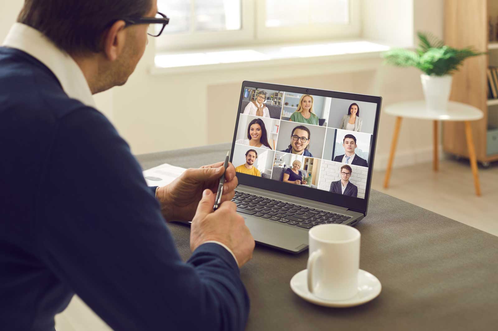 A person sits at a table, viewing a video call meeting on their laptop with several participants.