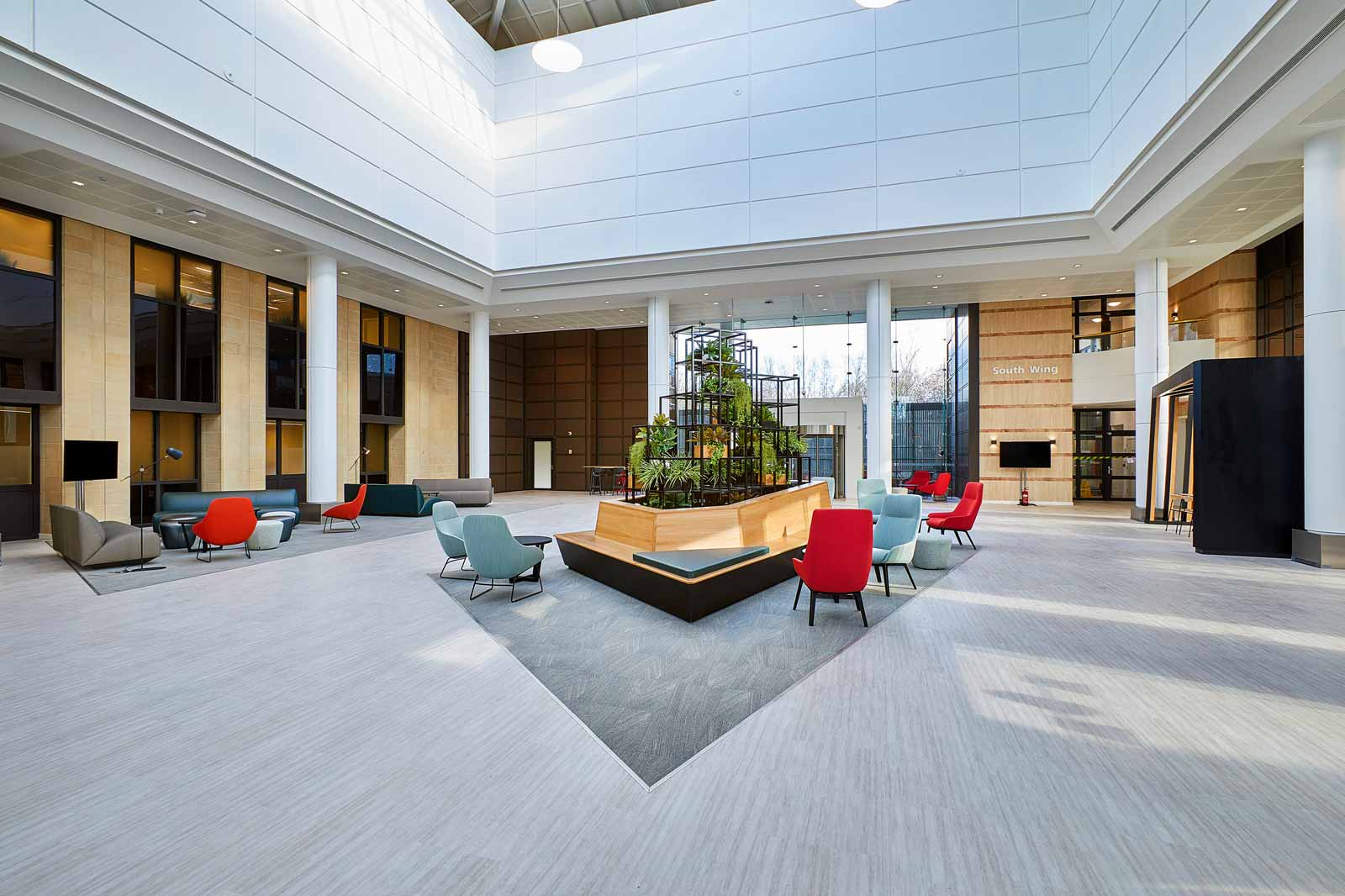 A bright indoor space featuring modern seating areas and a central plant installation. Large windows enhance natural light.