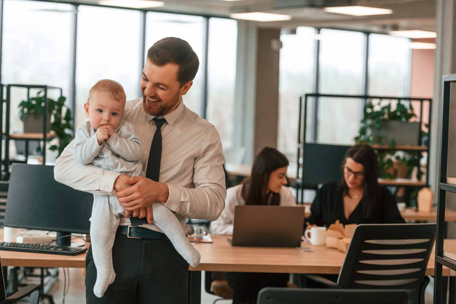 A man in a shirt and tie holds a baby. In the background, two women work on laptops in a modern office space.