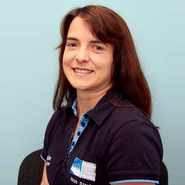 A woman with long brown hair, wearing a navy blue polo shirt with a logo, seated against a light blue background. Portrait of  Chedes Sarrionandia.