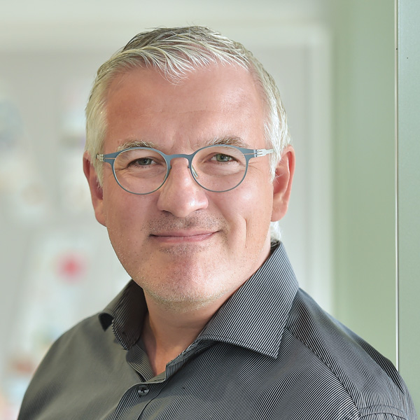 A man with grey hair wears a black striped shirt and stands in a bright room. Portrtait of Guido Hommelsheim.
