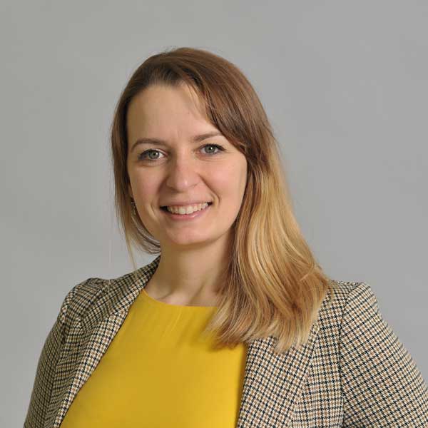 A person wearing a checkered blazer and a yellow top against a grey background. Portrtait of Ivana Rosenboom.