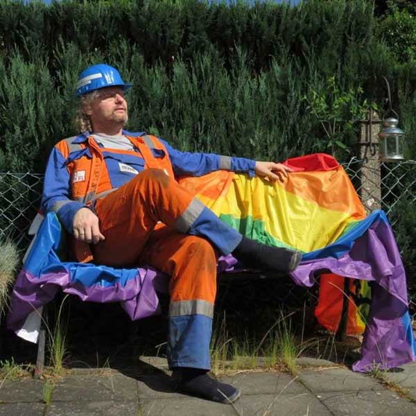 Eine Person in Arbeitskleidung sitzt entspannt auf einer Bank, umgeben von einem Regenbogenflaggen. Portrtait von Lothar Emmerich.