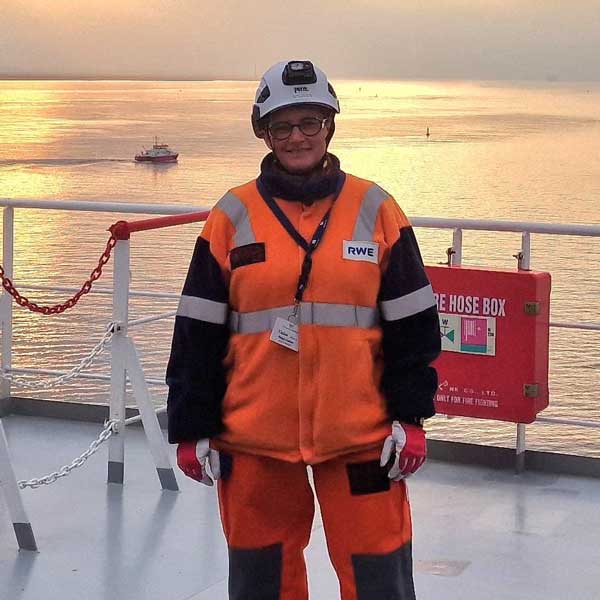 Eine Person in orangefarbener Arbeitskleidung steht auf einem Schiff mit Blick auf das Wasser und Sonnenuntergang. Portrtait von Malaika van Melick.