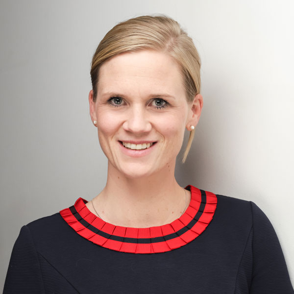 A woman wearing a navy dress with a red pleated collar, standing against a light grey background. Portrtait of Marie-Cecil Aufmkolk.
