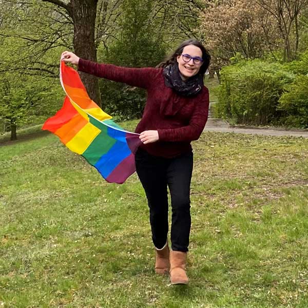 Eine Person in einem weinroten Pullover und einer schwarzen Hose, die eine Regenbogenflagge in einem grünen Park schwenkt. Portrait von Michaela Elschner.