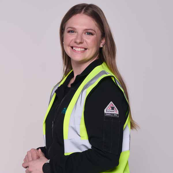 Eine Person trägt eine reflektierende Sicherheitsweste mit einem schwarzen Oberteil. Sie hat lange, braune Haare. Portrait von Samantha Mullin.