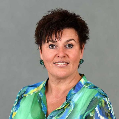 A person with short dark hair is wearing a colourful blouse and earrings. The background is grey. Portrait of Sandra Bossemeyer.