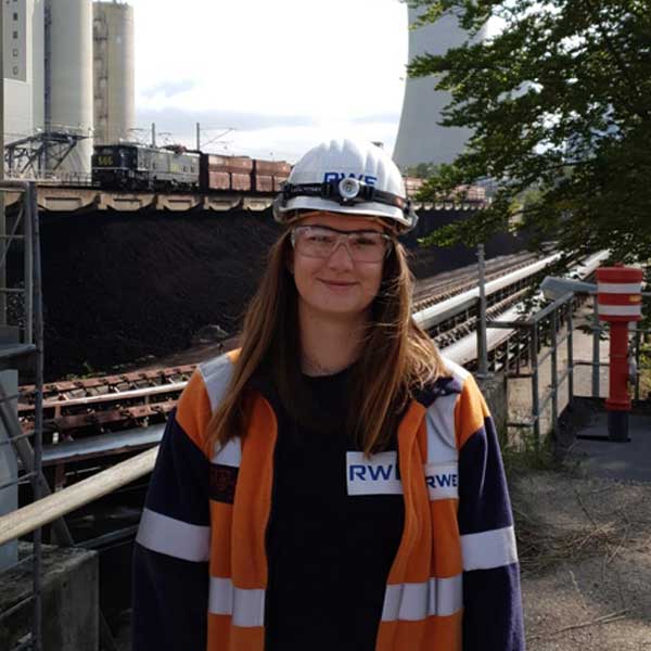 Eine Person mit Bauhelm und Sicherheitsjacke steht vor einer Kohlenlagerstätte mit Zügen im Hintergrund. Portrait von Sophia Lambertz.
