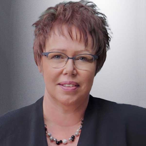 A person with short brown hair wears a black blouse and a necklace made of various beads. Portrait of Ulrike Pugh.