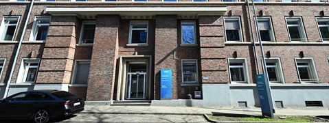 The image shows a modern brick building with windows and a sign in front of the entrance.
